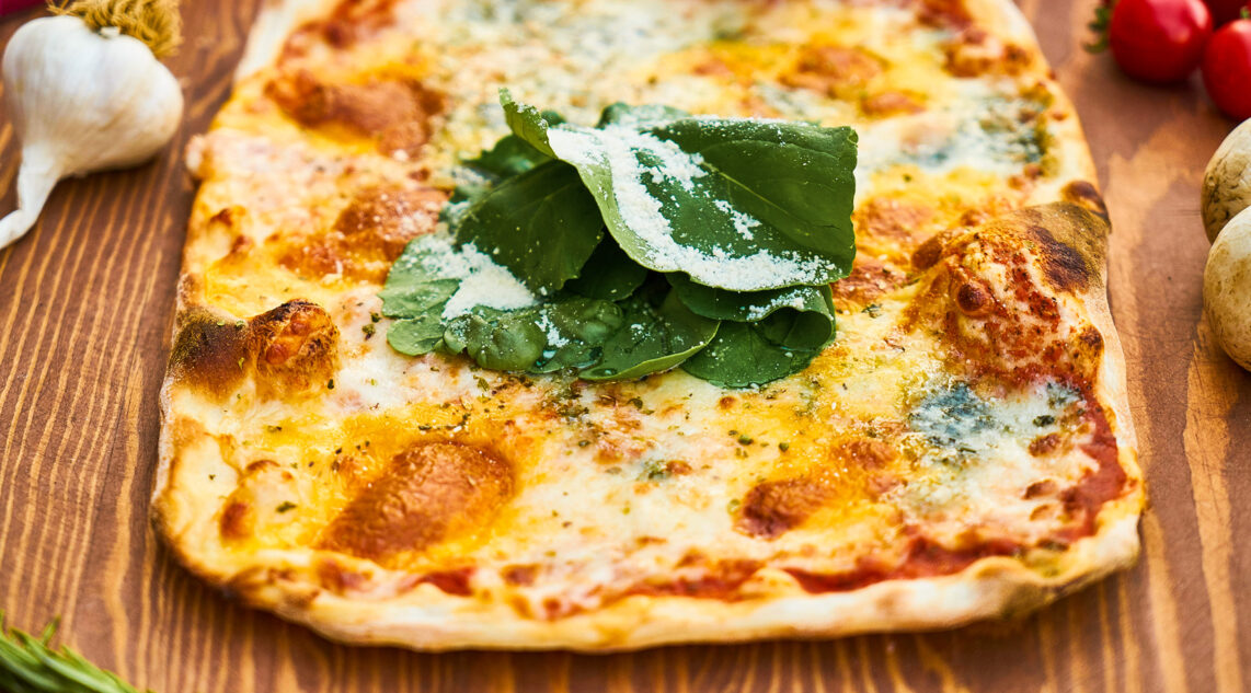 eine glutenfreie Pizza liegt fertig gebacken auf einem Holzbrett, sie ist Parmesan und Basilikum garniert, daneben liegen Tomaten, Knoblauch und Pilze