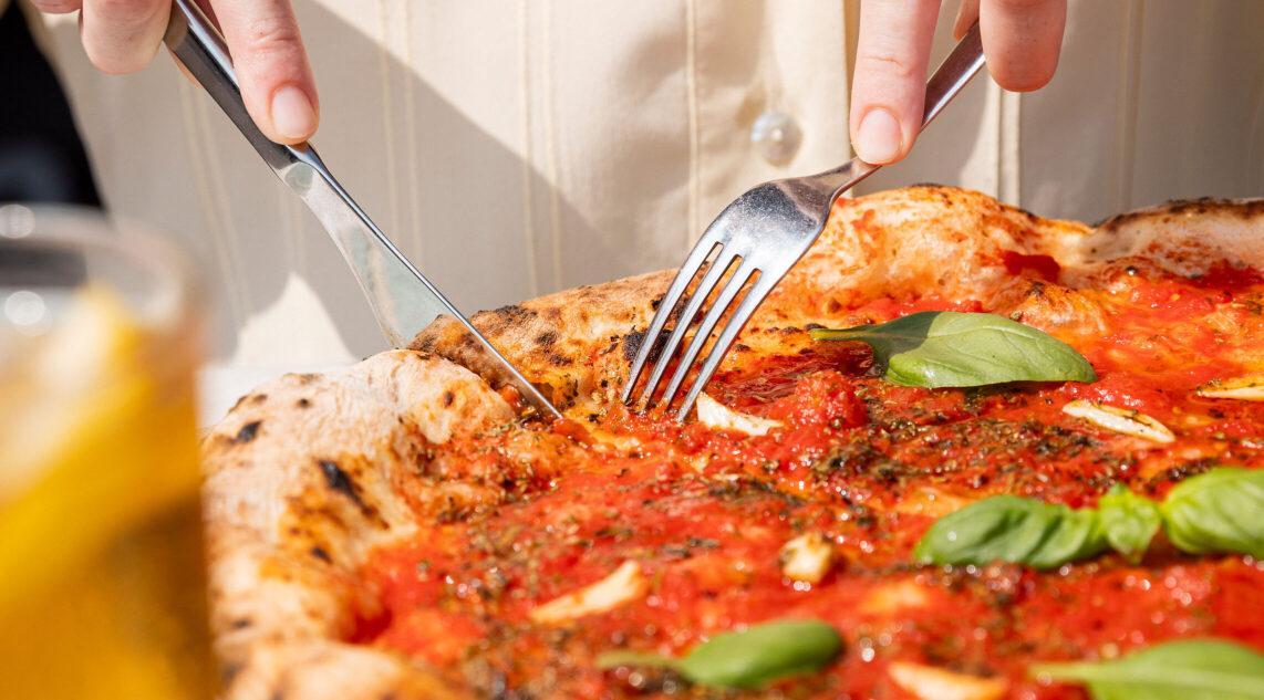 Nahaufnahme: in der Mittagssonne Neapels wird eine frisch gebackene Pizza Marinara mit Messer und Gabel geschnitten