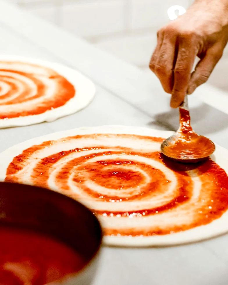 auf einer weißen Arbeitsfläche werden zwei Pizzateige in kreisförmigen Bewegungen mit Soße bestrichen