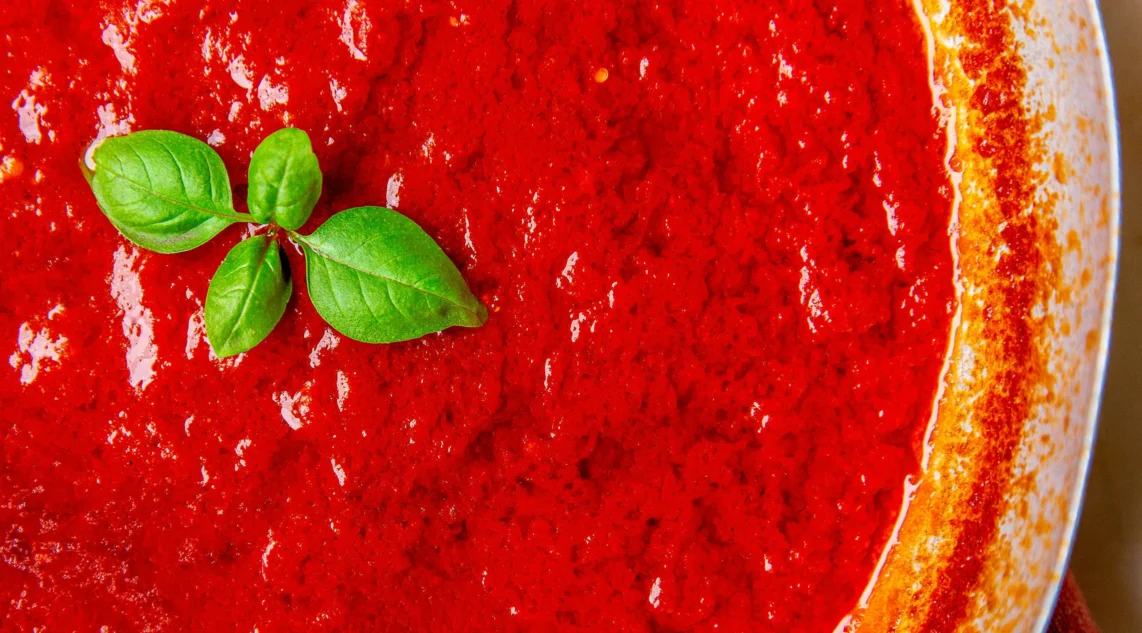 Nahaufnahme einer Metallpfanne in der frisch gekochtes klassisches Sugo zu erkennen ist, als Deko wurde die Spitze einer Basilikumpflanze auf die Soße platziert