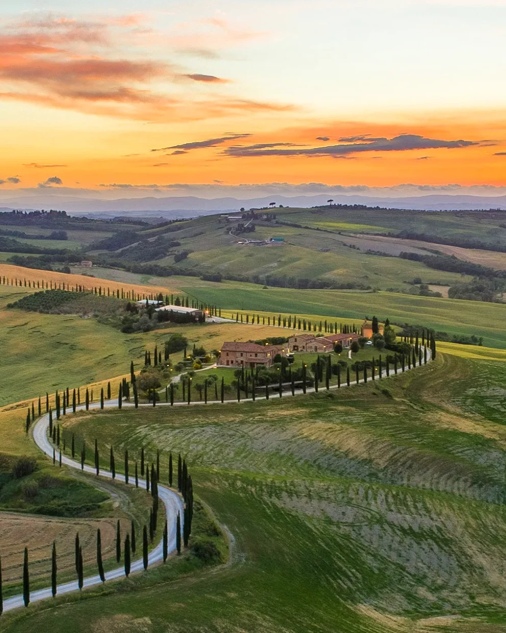 Sonnenuntergang im Panoramaformat: Das Valle D'Orcia mit grünen Hügeln, Zypressenbäumen und mediterranen Häusern liegt friedlich unter einem dramatischen Himmel in Orange-Blau-Violett