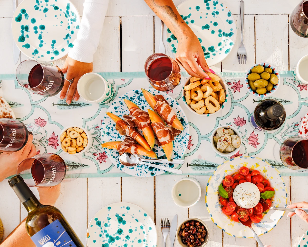 Draufsicht: ein reichlich gedeckter Tisch mit verschiedenen Antipasti auf einem tischläufer ist zu sehen, mehrer Personen sitzen am Tisch, eine Hand gießt Wein ein, eine andere greift gerade einen taralli