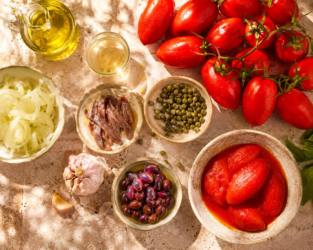Draufsicht auf sonnengereifte Tomaten, Sardellenfilets, Kapern, Taggiasca-Oliven, Knoblauch, Zwiebeln, Olivenöl und Weißwein.