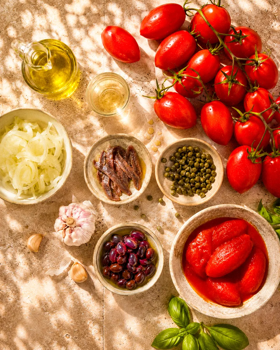 Draufsicht auf sonnengereifte Tomaten, Sardellenfilets, Kapern, Taggiasca-Oliven, Knoblauch, Zwiebeln, Olivenöl und Weißwein.