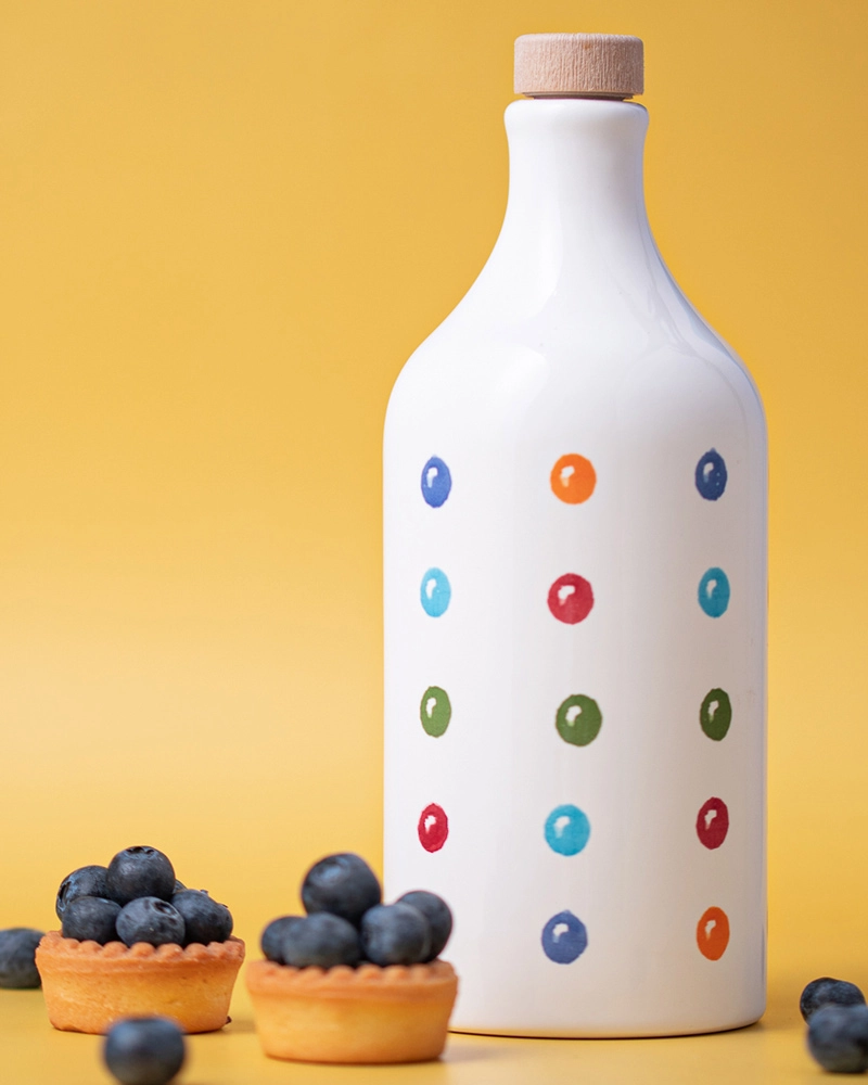 Muraglia Bouteille en terre cuite - Pois Muraglia Olivenöl in gepunkteter Keramikflasche vor gelbem Hintergrund, Blaubeer-Törtchen im Vordergrund