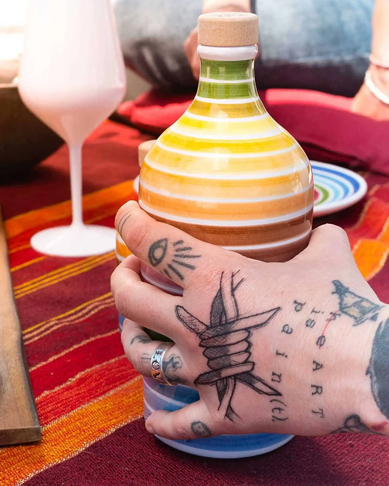 Muraglia Olivenöl - Regenbogen Keramikflasche – Flasche im Hintergrund, in bunter Keramikflasche präsentiert