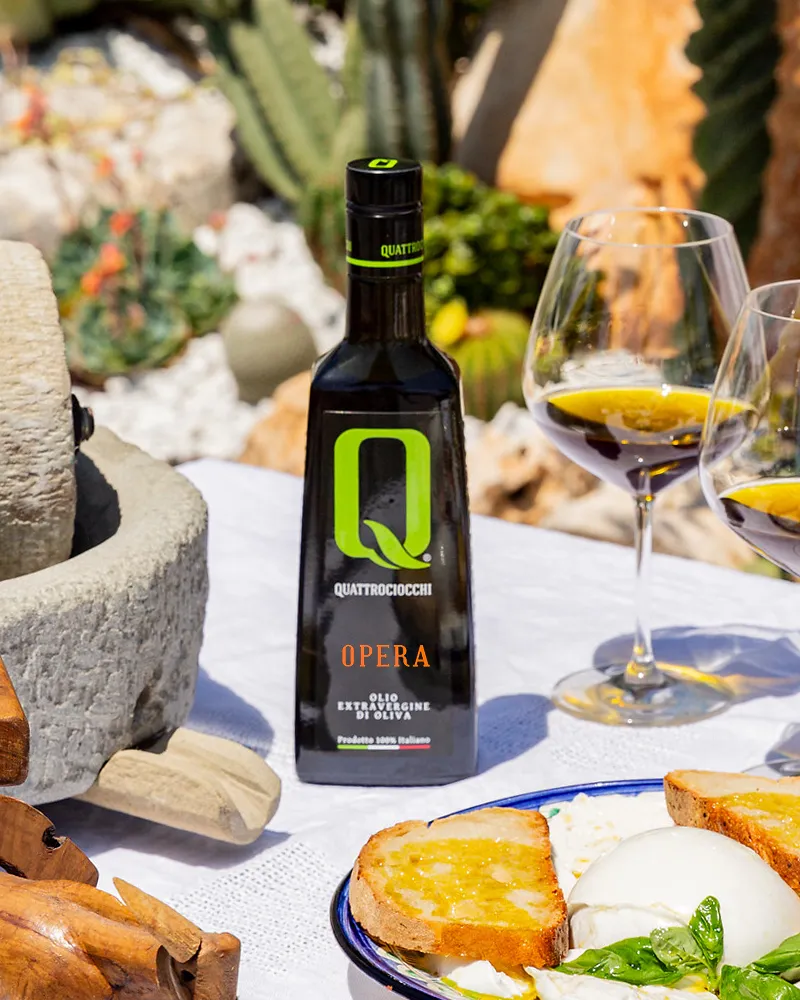 bestes Olivenöl 2025 Opera Flasche Quattrociocchi Olivenöl Extra Vergine 'Opera' neben zwei Gläsern mit Olivenöl und geröstetem Brot mit Mozzarella auf weißem Tischtuch im Freien
