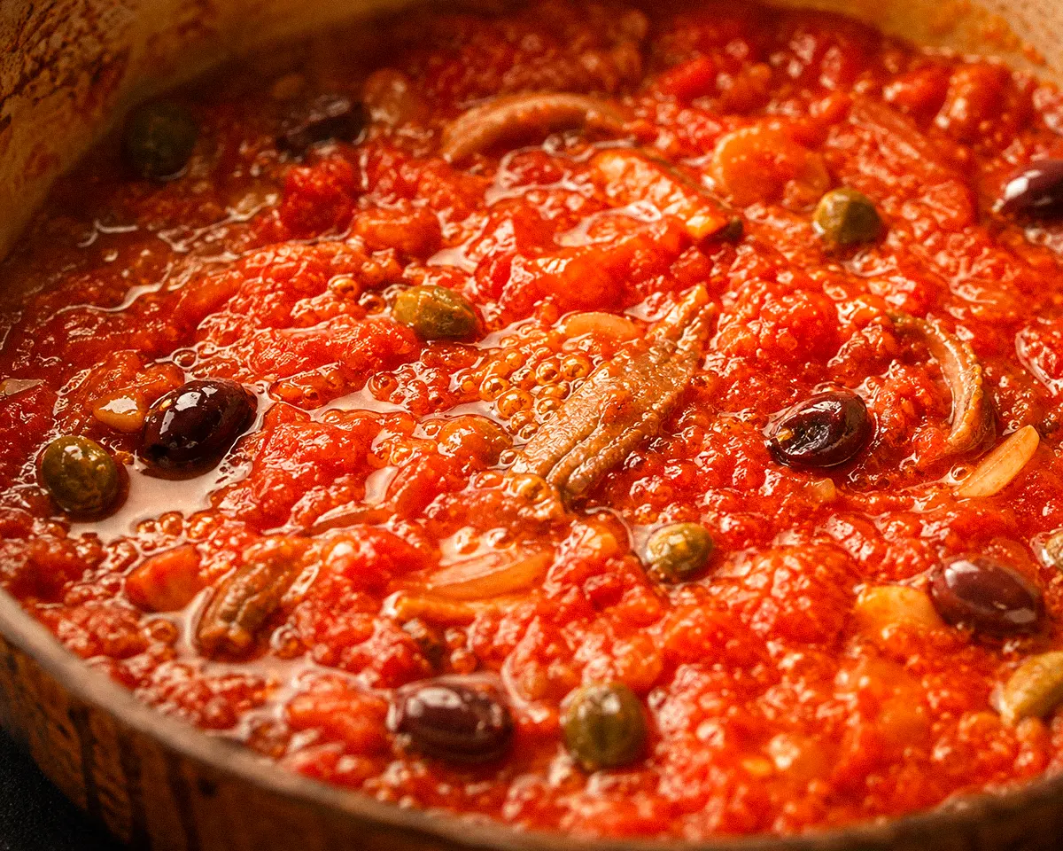 Nahaufnahme eines langsam köchelnden Tomatensugos mit Sardellen, Kapern, Oliven, Zwiebeln und Knoblauch.